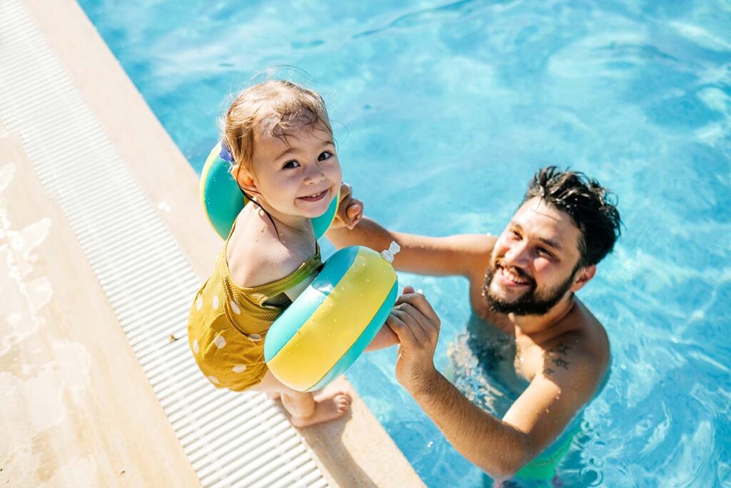 child wearing floaties standing on the edge of a pool; an adult in the pool is holding her hands