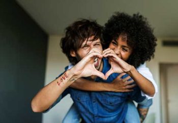 A man and woman are forming a heart shape with their hands. She is on his back. They're smiling.