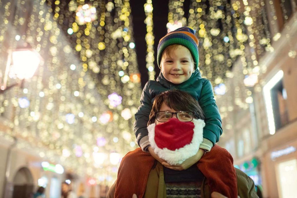 a parent wearing a holiday face mask, holding child on shoulders in front of street scape decorated for the holidays