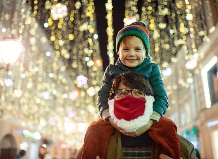 a parent wearing a holiday face mask, holding child on shoulders in front of street scape decorated for the holidays