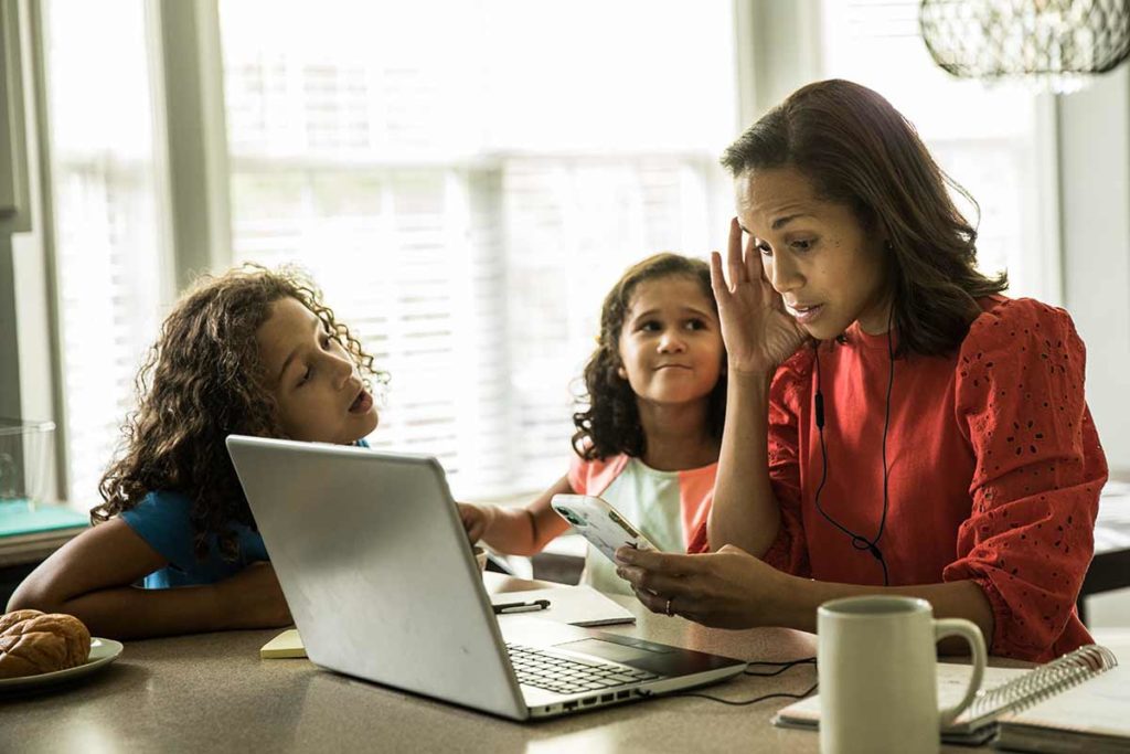 woman at home sitting in front of laptop with two kids trying to get her attention. she's feeling the effects of stress