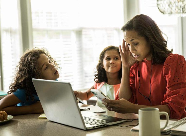 woman at home sitting in front of laptop with two kids trying to get her attention. she's feeling the effects of stress