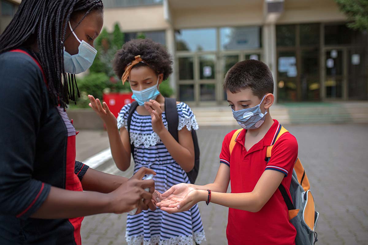 two kids wearing masks and backpacks, standing outside a school, getting hand sanitizer from adult