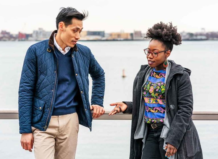 A man and a woman standing by a railing in front of water discussing things not to say to someone with a chronic illness.