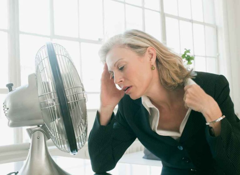 A woman sitting in front of a fan because she is having hot flashes from perimenopause.