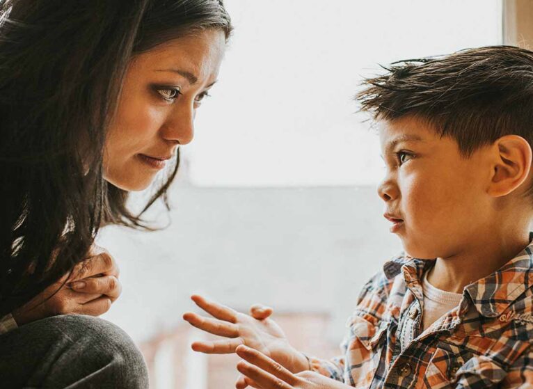 A cute young boy gestures using his hands. A woman, who sits close-by, listens and watches him intently as he explains something.