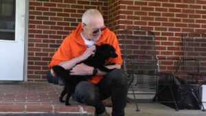 Jerry Austin wearing an orange shirt and dark pants, sitting on a porch and holding a black dog. He opted for a lasting open aortic aneurysm repair when it came time for surgery.