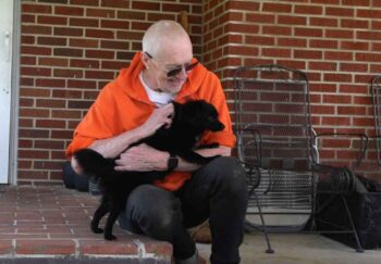 Jerry Austin wearing an orange shirt and dark pants, sitting on a porch and holding a black dog. He opted for a lasting open aortic aneurysm repair when it came time for surgery.