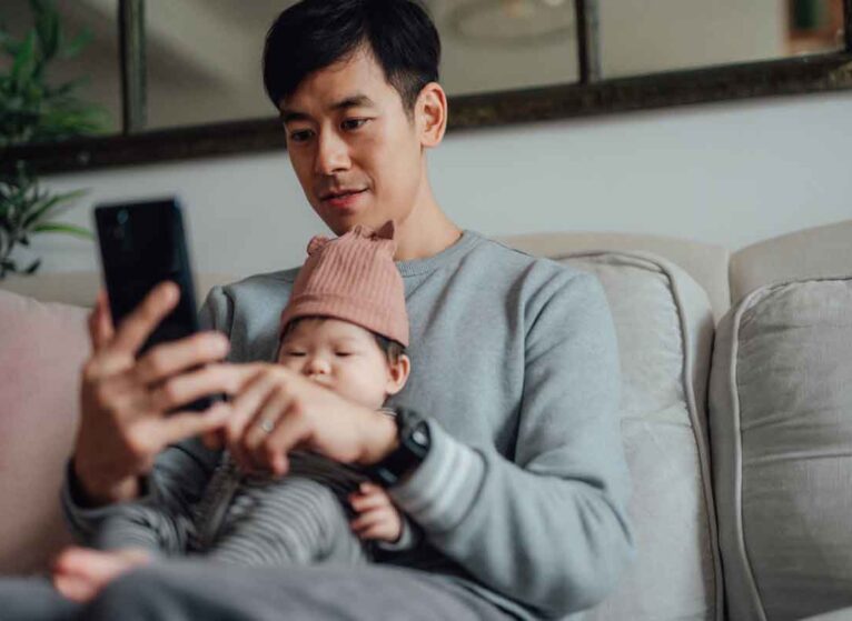 A man sitting while holding a baby and looking at his phone