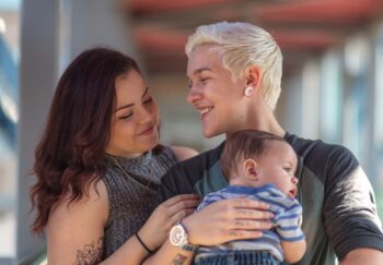 Portrait of a happy LGBT family. The young adult partners are spending time with their baby. The non-binary gendered adult is holding the baby boy. The parents are smiling.