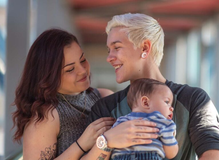 Portrait of a happy LGBT family. The young adult partners are spending time with their baby. The non-binary gendered adult is holding the baby boy. The parents are smiling.