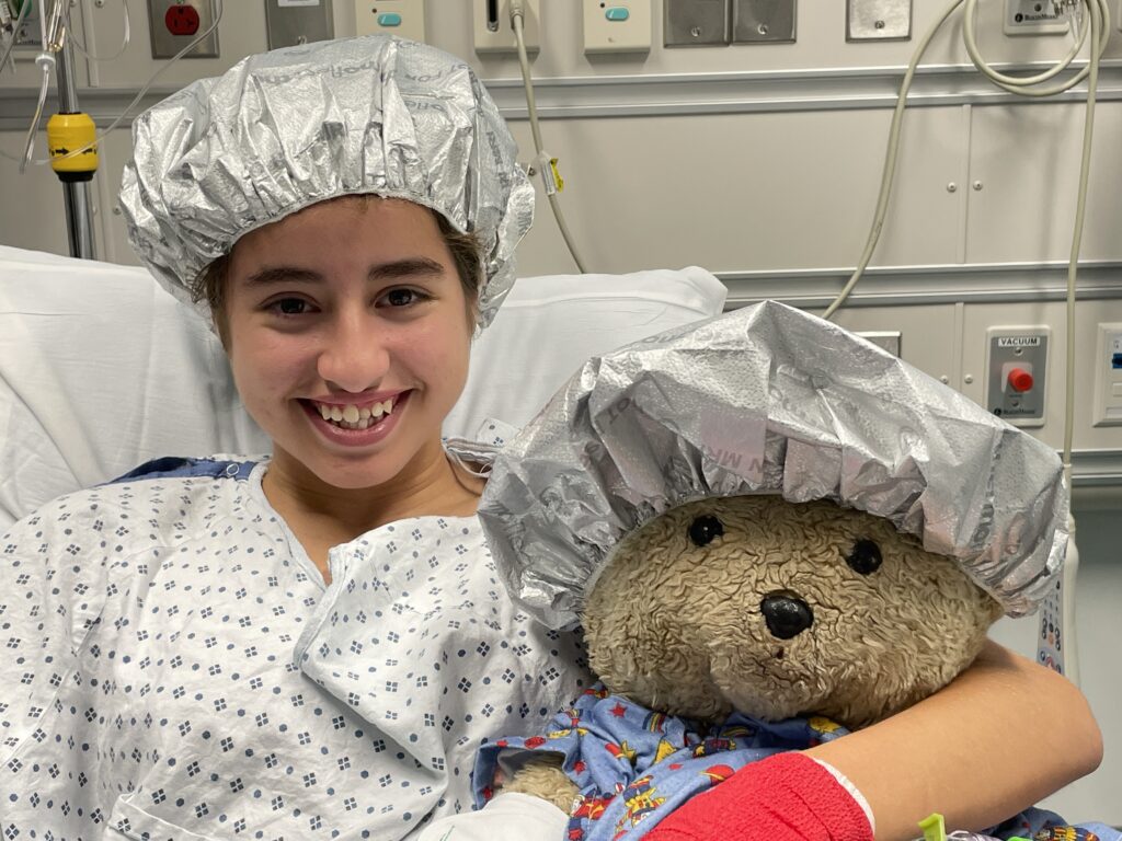 Olivia smiles at camera before surgery with her hair in a surgical cap. She holds up a teddy bear also wearing a surgical cap.