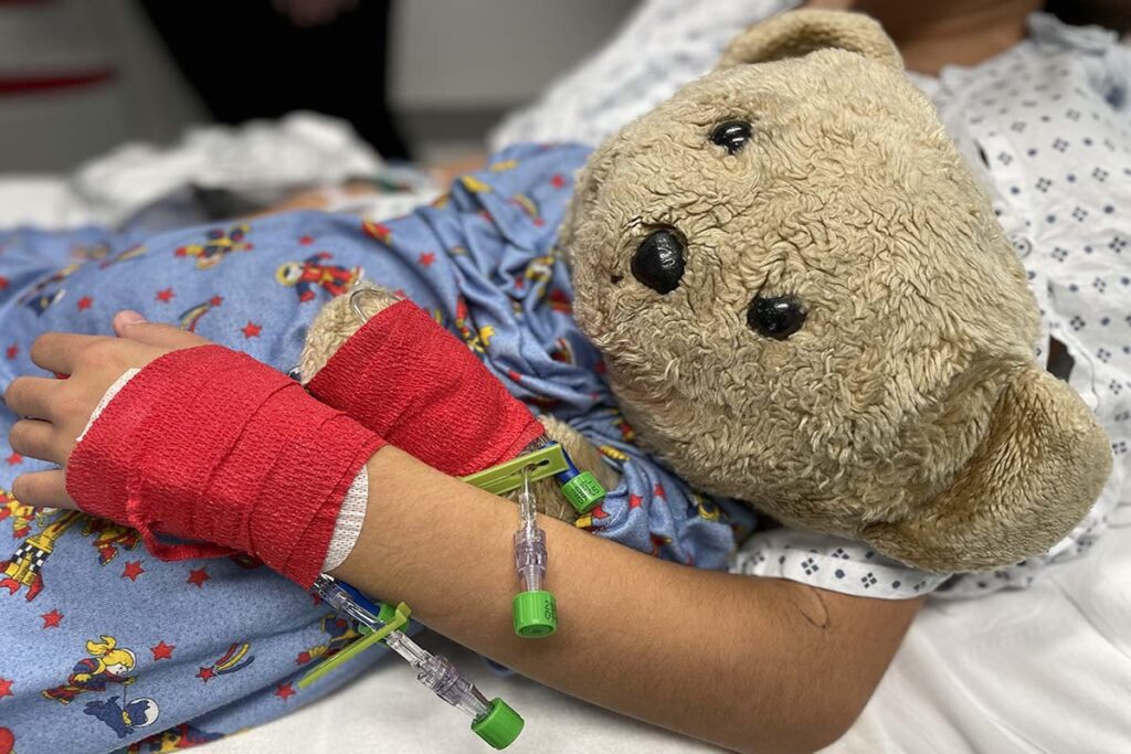 Young girl's arm with IV and wrap hugs a teddy bear before surgery.