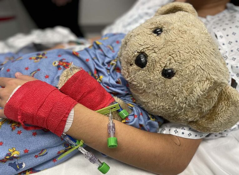 Young girl's arm with IV and wrap hugs a teddy bear before surgery.