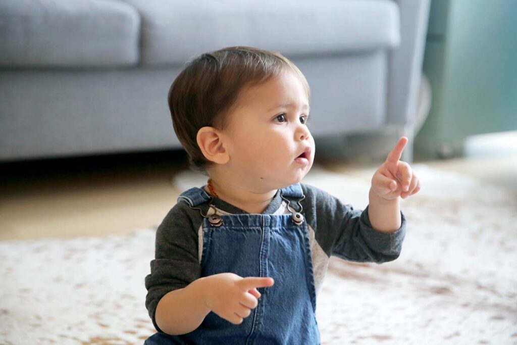 A LatinX toddler points his index finger, while sitting in his living room wearing jean overalls and a long sleeved t-shirt.