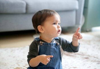 A LatinX toddler points his index finger, while sitting in his living room wearing jean overalls and a long sleeved t-shirt.