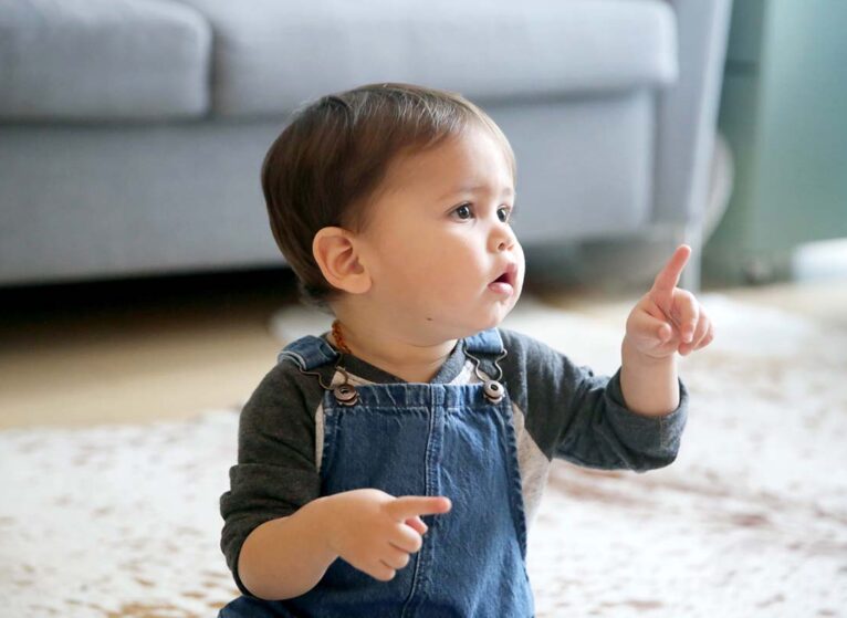 A LatinX toddler points his index finger, while sitting in his living room wearing jean overalls and a long sleeved t-shirt.