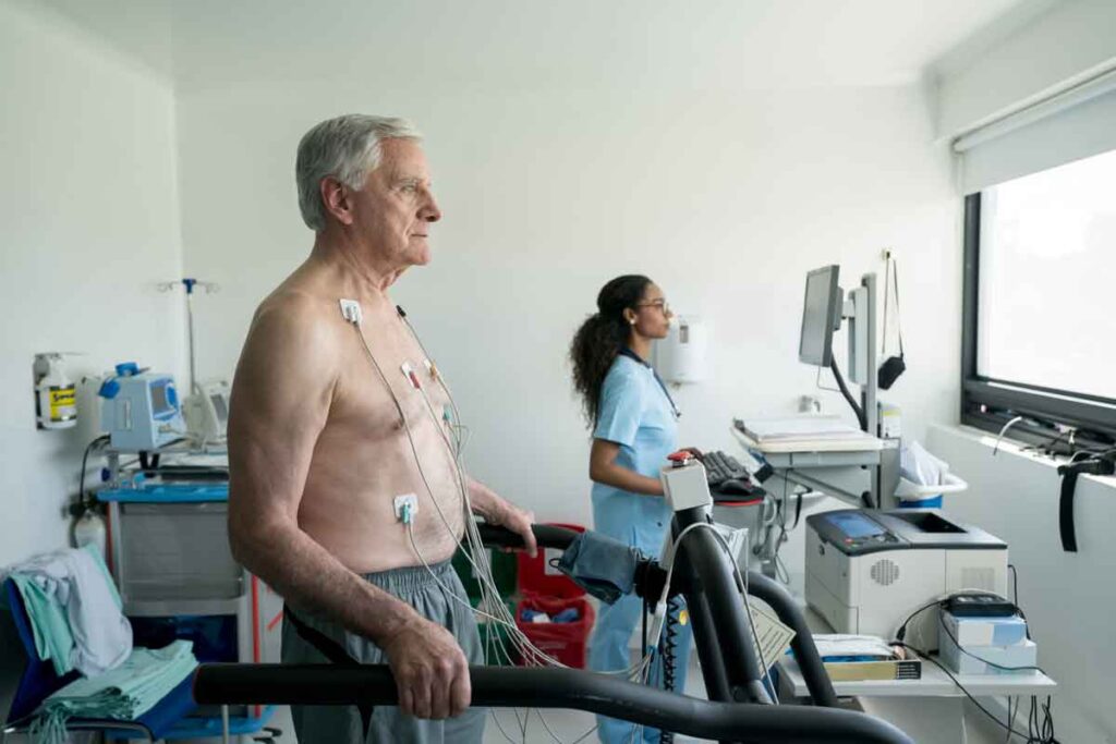 A man having heart tests, he has electrodes on his chest looking to the right while a healthcare provider looks at a screen in the background