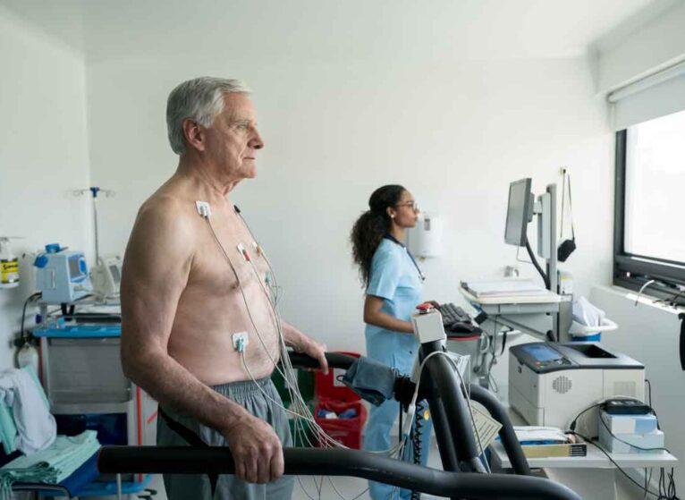 A man having heart tests, he has electrodes on his chest looking to the right while a healthcare provider looks at a screen in the background