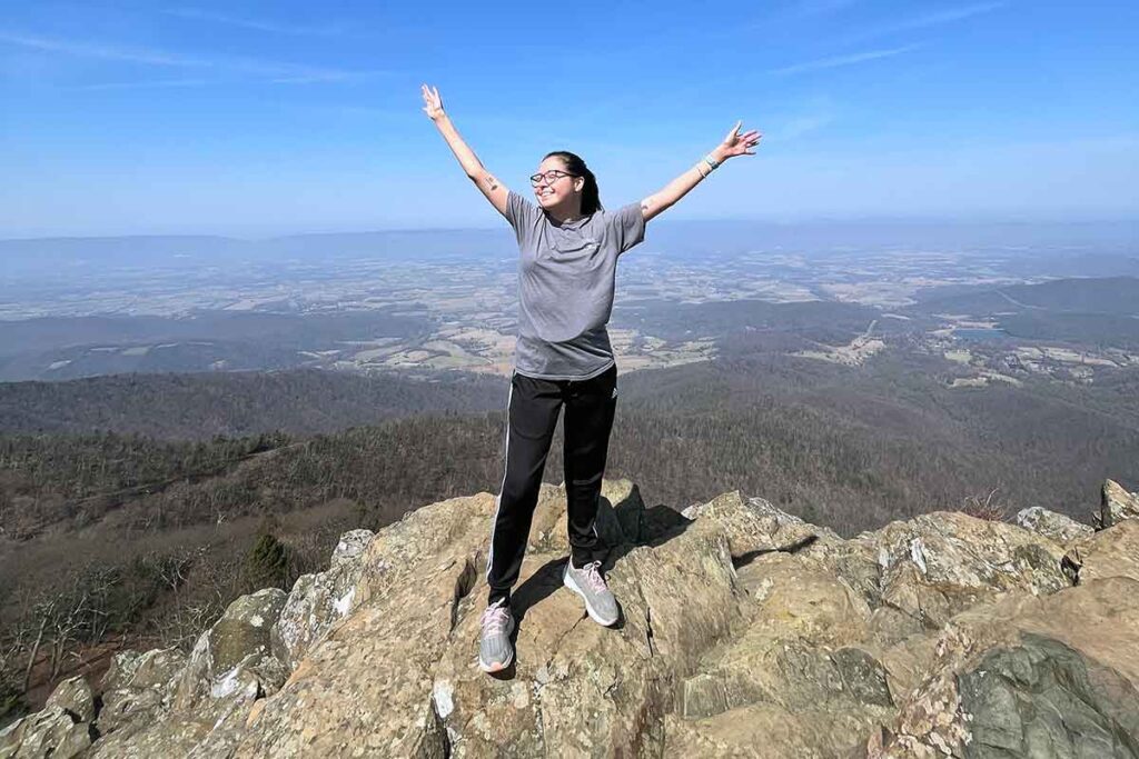 A young transplant survivor celebrates with arms out on top of a mountain.