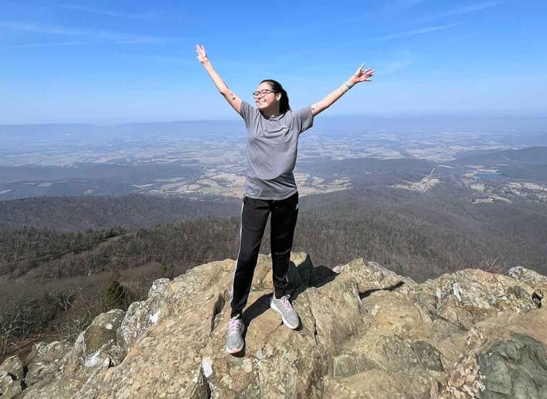 A young transplant survivor celebrates with arms out on top of a mountain.
