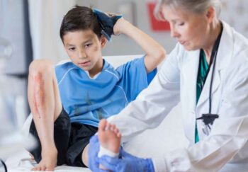 A young boy holding an ice pack to his head, with a cut on his shin and getting his ankle wrapped by a healthcare [rovider