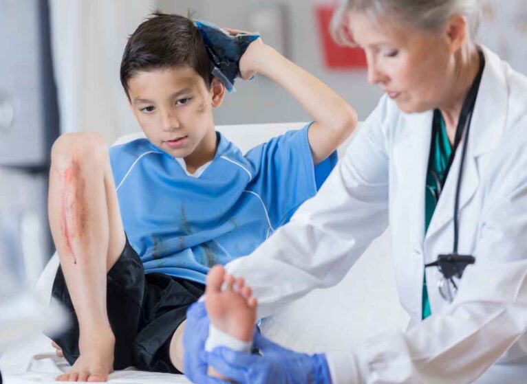 A young boy holding an ice pack to his head, with a cut on his shin and getting his ankle wrapped by a healthcare [rovider