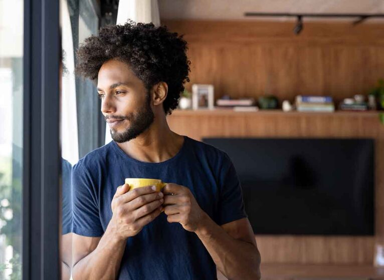 A man looking out the window with a mug of warm water in his hands