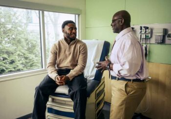 A young man sitting in a medical office talking about a medical device recall with a doctor who is standing