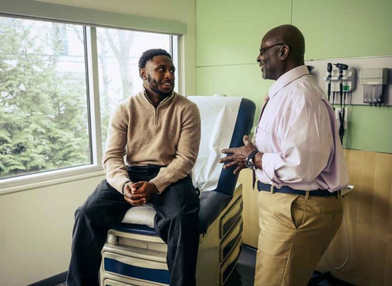 A young man sitting in a medical office talking about a medical device recall with a doctor who is standing