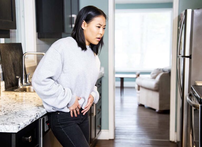 A woman holding her abdominal region in pain standing in her kitchen